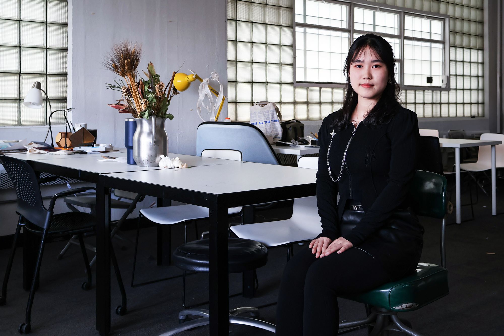 Young woman seated in a studio or classroom space, dressed in black with a long necklace. She sits beside a table decorated with a silver vase containing dried plants and flowers. The room features glass block windows, various chairs, and scattered design materials.